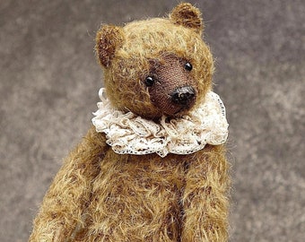 Puede incluir: Un pequeño oso de peluche marrón con un collar de encaje blanco. El oso está sentado en una mano, y sus patas son visibles.