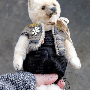 May include: A cream-colored teddy bear with dark eyes and a dark nose, wearing a gray and brown striped knit vest with a white snowflake-shaped button, a beige ribbon around its neck, and black textured overalls. Text reads Tiny Hearts Gerlinn Bears.