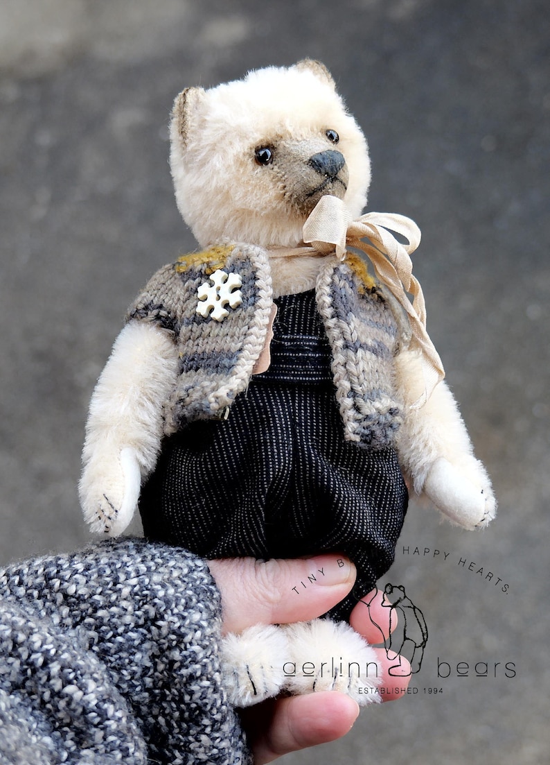 May include: A cream-colored teddy bear with dark eyes and a dark nose, wearing a gray and brown striped knit vest with a white snowflake-shaped button, a beige ribbon around its neck, and black textured overalls. Text reads Tiny Hearts Gerlinn Bears.