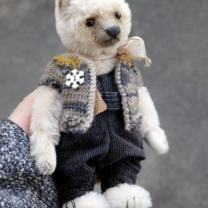May include: A small, cream-colored plush bear with dark eyes and a dark nose. The bear wears a grey and mustard-yellow knitted vest featuring a white snowflake-shaped button, along with dark grey pinstriped overalls and a light beige ribbon neck tie.