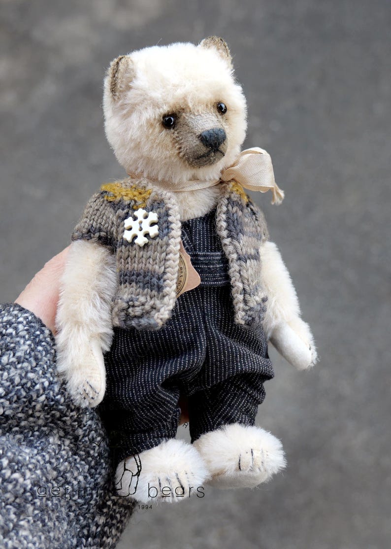 May include: A small, cream-colored plush bear with dark eyes and a dark nose. The bear wears a grey and mustard-yellow knitted vest featuring a white snowflake-shaped button, along with dark grey pinstriped overalls and a light beige ribbon neck tie.