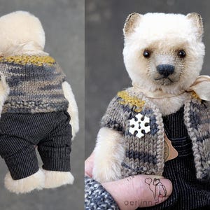 May include: A plush bear with cream-colored fur wearing a knitted vest in shades of gray and mustard yellow, dark pinstriped trousers, and a white snowflake-shaped pin. A light beige ribbon is tied around the neck of the bear.