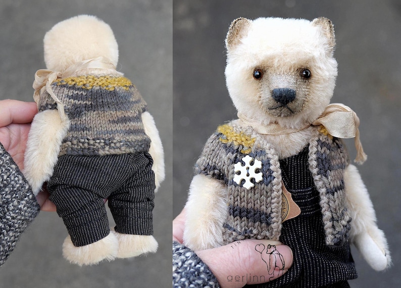 May include: A plush bear with cream-colored fur wearing a knitted vest in shades of gray and mustard yellow, dark pinstriped trousers, and a white snowflake-shaped pin. A light beige ribbon is tied around the neck of the bear.