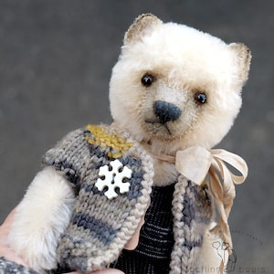 May include: A cream-colored teddy bear with dark eyes and a black nose, wearing a grey and brown knit vest featuring a white snowflake-shaped button and a beige ribbon tied around its neck. The bear has a logo with the text BEARS HAPPY HEARTS.