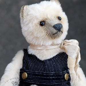 May include: A plush bear with cream-colored fur, dark eyes, and a dark nose, wearing dark blue pinstriped overalls with brass-colored buttons and a light beige ribbon tied around its neck. The bear features a logo with the text Tiny Bears.