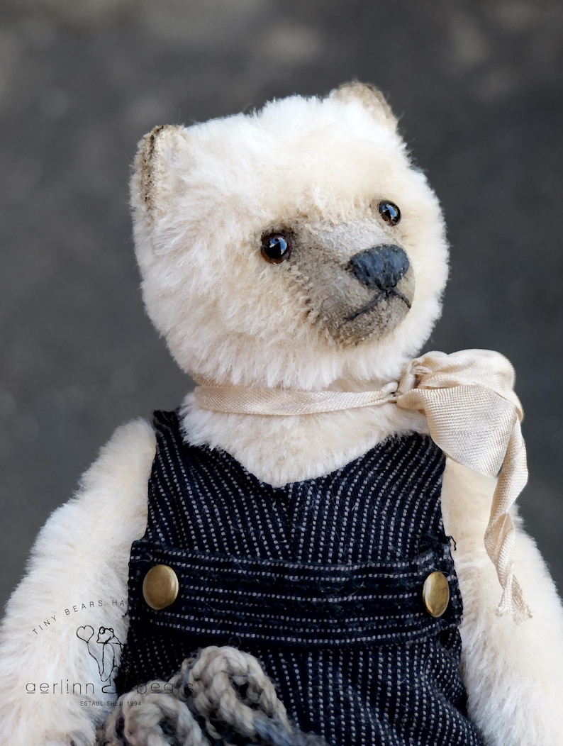 May include: A plush bear with cream-colored fur, dark eyes, and a dark nose, wearing dark blue pinstriped overalls with brass-colored buttons and a light beige ribbon tied around its neck. The bear features a logo with the text Tiny Bears.