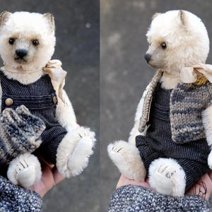 May include: A small, cream-colored plush bear with dark eyes and a stitched nose. The bear wears dark gray corduroy overalls with brass buttons, a light beige ribbon tied around its neck, and a multi-colored knitted vest with gray and brown tones.