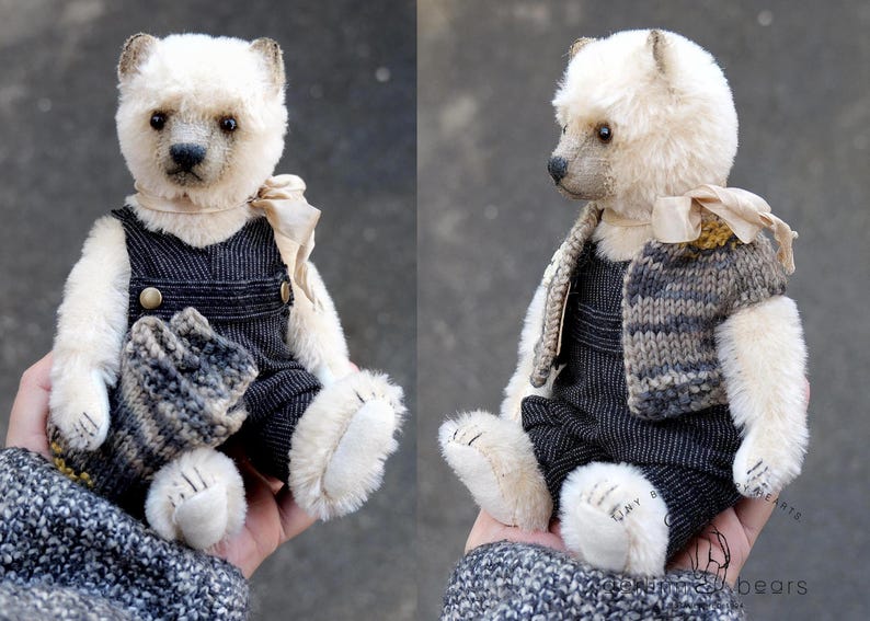 May include: A small, cream-colored plush bear with dark eyes and a stitched nose. The bear wears dark gray corduroy overalls with brass buttons, a light beige ribbon tied around its neck, and a multi-colored knitted vest with gray and brown tones.