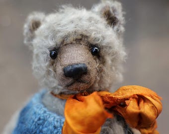 May include: A handmade teddy bear with a gray, fluffy fur and a brown muzzle. The bear wears a blue sweater and an orange scarf tied around its neck. The bear has black eyes.