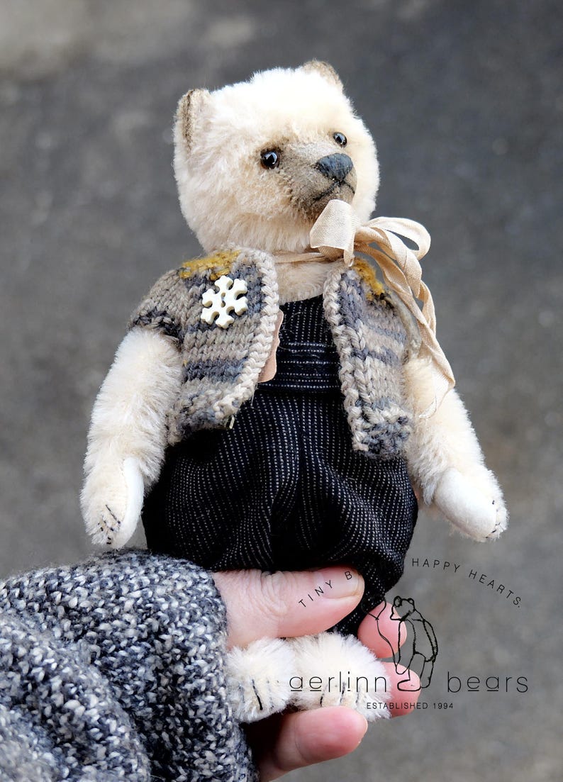 May include: A cream-colored teddy bear with dark eyes and a dark nose, wearing a gray and brown striped knit vest with a white snowflake-shaped button, a beige ribbon around its neck, and black textured overalls. Text reads Tiny Hearts Gerlinn Bears.