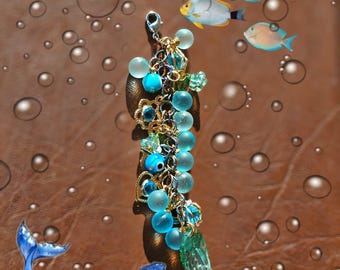 May include: A blue and green charm bracelet with a whale charm, blue glass beads, and gold accents. The bracelet is surrounded by bubbles and fish.
