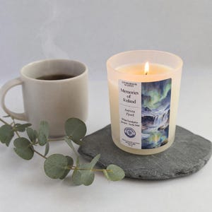 May include: A lit candle in a frosted glass jar with a label reading Storybook Scents Memories of Iceland Aurora Fjord White Eucalyptus Juniper Arctic Mist 150 g. Next to it is a steaming ceramic mug and a sprig of eucalyptus on a slate coaster.