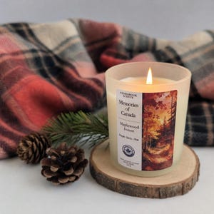 May include: A lit candle in a frosted glass jar with a label reading Storybook Scents Memories of Canada Maplewood Forest Maple Birch Pine 150 g. The candle sits on a round wood slice next to two pinecones and a plaid fabric.
