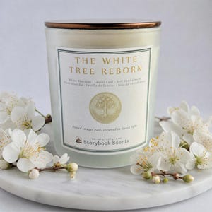 May include: A scented candle in a frosted glass jar with a bronze lid. The label reads THE WHITE TREE REBORN, White Blossom, Laurel Leaf, Soft Sandalwood, and Storybook Scents. It notes 40-50 h, 227 g, 8 oz. White flowers surround the base of the jar.