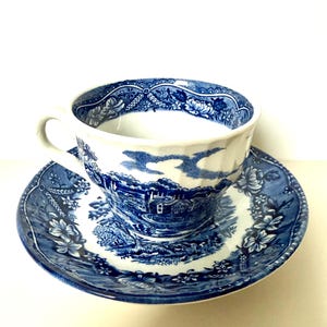 Op de afbeelding: Een blauw-wit kop en schotel set. De kop en schotel hebben een gedetailleerd landschapsontwerp met bloemen- en geometrische patronen. De kop heeft een gebogen oor en staat op een bijpassende schotel.