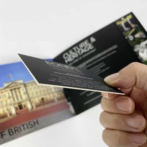 May include: A black business card with white text is held in front of a brochure. The card reads "BONDS LONDON" and includes contact information. The brochure features a photo of a building and the words "OF BRITISH" and "CULTURE & HERITAGE".