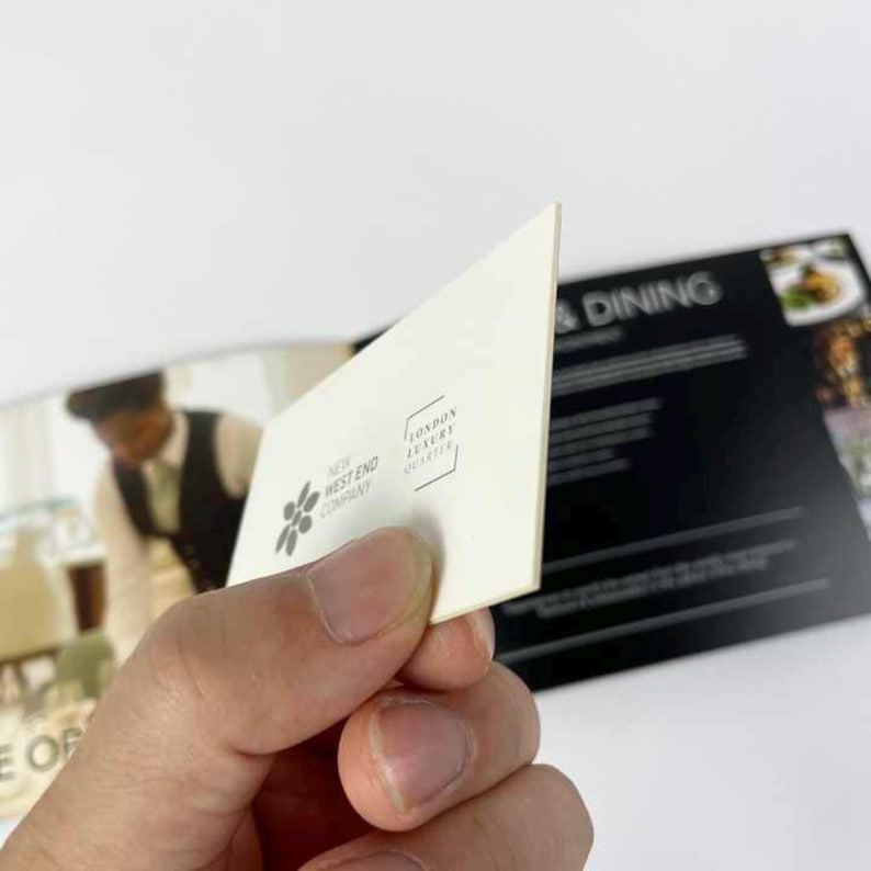 May include: A cream-colored business card held in a hand, featuring the text "NEW WEST END COMPANY" and a floral logo. The card also displays "LONDON LUXURY QUARTER" in a rectangular frame. The background shows a restaurant menu with the word "DINING".