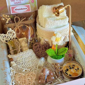 May include: A gift box filled with various items, including a small white towel, a candle, a small potted plant, and a bag of dried oranges. The box also contains a card that reads "THE PERFECKT TIME TO MAKE WISHES" and a sticker that says "Abundance".