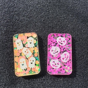 May include: Two rectangular Halloween resin decorations. One has white ghost designs on orange and yellow glitter. The other has white jack-o'-lantern designs on a pink glitter background. Both have a clear resin finish.