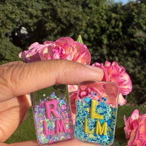 May include: Two rectangular resin pendants, each with glitter and letters. One pendant has pink letters "R L M" on a background of pink, purple, and green glitter. The other has yellow letters "L L M" on a blue glitter background.