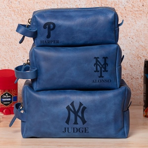 May include: Three stacked blue leather toiletry bags with personalized initials and names: "P HARPER", "M ALONSO", and "NY JUDGE". The bags are on a wooden surface with grooming products.