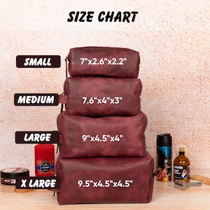 Op de afbeelding: Een maattabel toont vier bordeauxrode leren toilettassen in verschillende maten: klein (17.8cm x 6.6cm x 5.6cm), medium (19.3cm x 10.2cm x 7.6cm), groot (22.9cm x 11.4cm x 10.2cm) en extra groot (24.1cm x 11.4cm x 11.4cm). De tekst "SIZE CHART" staat bovenaan.
