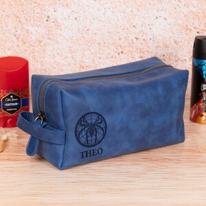 Op de afbeelding: Een blauwe leren toilettas met ritssluiting en handvat. De tas heeft een zwart spinnenwebontwerp met de naam "THEO" eronder. Andere verzorgingsproducten zijn zichtbaar op de achtergrond.