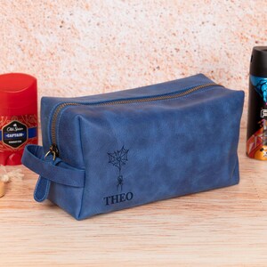 Op de afbeelding: Een blauwe leren toilettas met ritssluiting en handvat. De tas heeft een spinnenweb- en spinnenontwerp met de naam "THEO" eronder. Een rode deodorantstick en een zwart-blauwe spuitfles staan aan weerszijden.