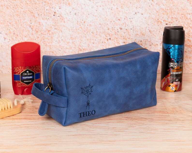 Op de afbeelding: Een blauwe leren toilettas met ritssluiting en handvat. De tas heeft een spinnenweb- en spinnenontwerp met de naam "THEO" eronder. Een rode deodorantstick en een zwart-blauwe spuitfles staan aan weerszijden.