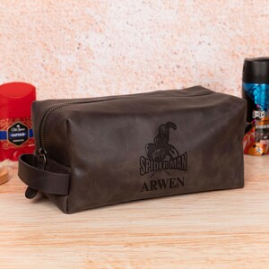 Op de afbeelding: Een donkerbruine leren toilettas met ritssluiting en een handvat. De tas heeft een Spider-Man-afbeelding en de naam "ARWEN" op de voorkant gedrukt. Andere verzorgingsproducten zijn zichtbaar op de achtergrond.