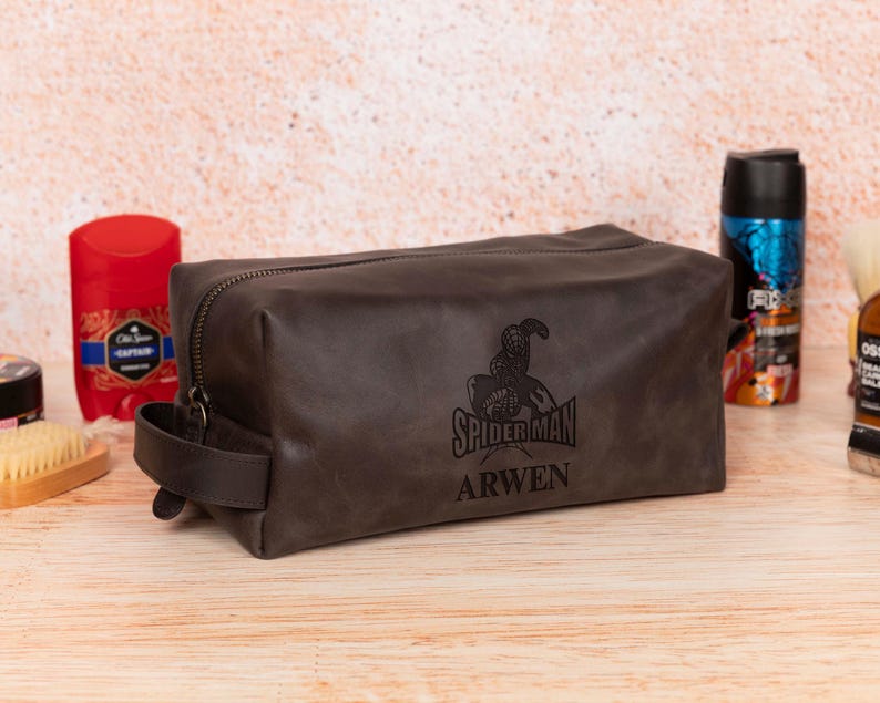 Op de afbeelding: Een donkerbruine leren toilettas met ritssluiting en een handvat. De tas heeft een Spider-Man-afbeelding en de naam "ARWEN" op de voorkant gedrukt. Andere verzorgingsproducten zijn zichtbaar op de achtergrond.