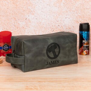 Op de afbeelding: Een grijze leren toilettas met ritssluiting en een handvat. De tas heeft een cirkelvormig embleem met een hamerontwerp en de naam "JAMES" eronder. Andere verzorgingsproducten zijn zichtbaar op de achtergrond.