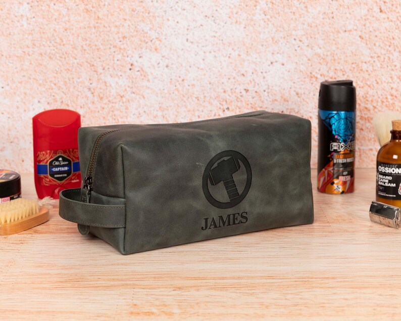 Op de afbeelding: Een grijze leren toilettas met ritssluiting en een handvat. De tas heeft een cirkelvormig embleem met een hamerontwerp en de naam "JAMES" eronder. Andere verzorgingsproducten zijn zichtbaar op de achtergrond.