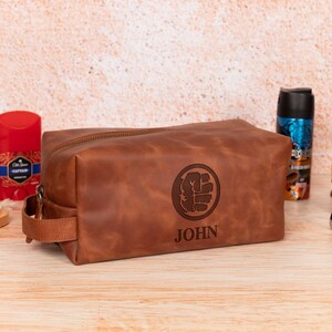Op de afbeelding: Een bruine leren toilettas met ritssluiting en handvat. De tas heeft een gegraveerd ontwerp van een vuist in een cirkel en de naam "JOHN" eronder. Andere verzorgingsproducten zijn zichtbaar op de achtergrond.