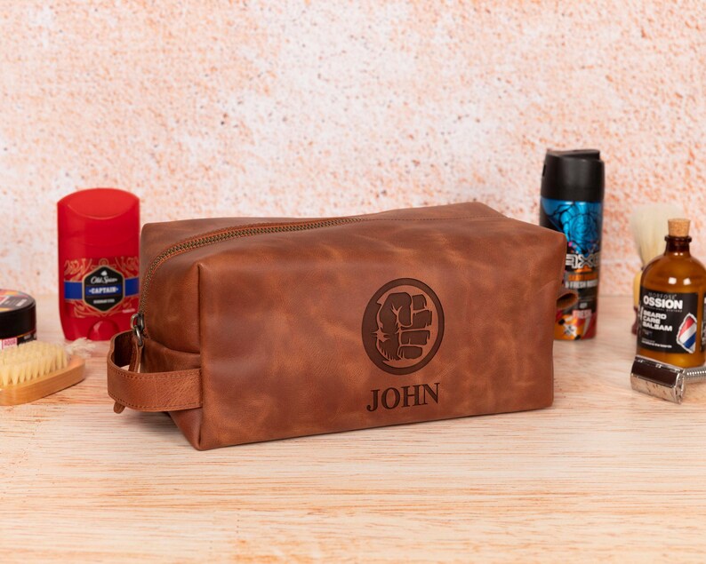 Op de afbeelding: Een bruine leren toilettas met ritssluiting en handvat. De tas heeft een gegraveerd ontwerp van een vuist in een cirkel en de naam "JOHN" eronder. Andere verzorgingsproducten zijn zichtbaar op de achtergrond.