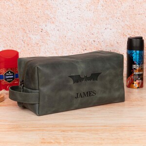 Op de afbeelding: Een donkergrijze leren toilettas met ritssluiting en een handvat. De tas heeft een Batman-logo en de naam "JAMES" gegraveerd op de voorkant. Andere verzorgingsproducten zijn zichtbaar op de achtergrond.
