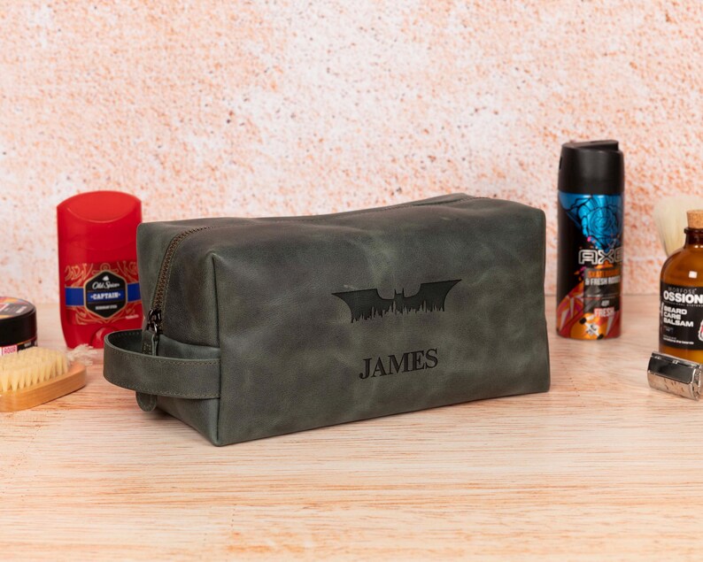 Op de afbeelding: Een donkergrijze leren toilettas met ritssluiting en een handvat. De tas heeft een Batman-logo en de naam "JAMES" gegraveerd op de voorkant. Andere verzorgingsproducten zijn zichtbaar op de achtergrond.