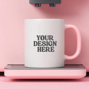 May include: White ceramic mug with a pink handle, featuring the text "YOUR DESIGN HERE" in a bold, dark font. The mug is placed on a pink surface, with a pink and black machine in the background. The overall aesthetic is clean and modern.