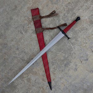 Könnte beinhalten: Ein langes, silbernes Schwert mit schwarzem Griff und Parierstange, neben einer roten Lederscheide. Die Scheide hat braune Lederriemen und ein gewebtes Muster. Die Klinge des Schwertes ist scharf und spitz.