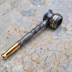 Könnte beinhalten: Ein handgefertigter Hammer mit gemustertem Stahlkopf und -griff, akzentuiert mit goldfarbenen Bändern und einer goldfarbenen Schlagfläche. Der Hammer wird auf einer strukturierten, grauen Oberfläche präsentiert.