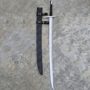 Puede incluir: Una espada larga y plateada con empuñadura y guarda negras, acompañada de una vaina de cuero negro. La espada tiene una hoja pulida y una punta. La vaina tiene dos correas con hebillas color latón.