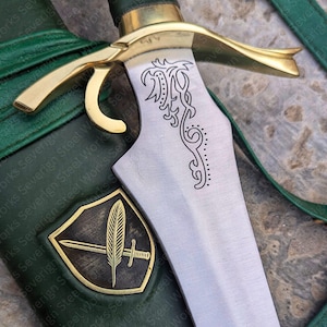 Puede incluir: Primer plano de una daga decorativa con una hoja plateada y una empuñadura dorada. La hoja presenta un diseño grabado. La daga está en una vaina de cuero verde con un emblema dorado. El texto "Severign Steel Works" es visible.