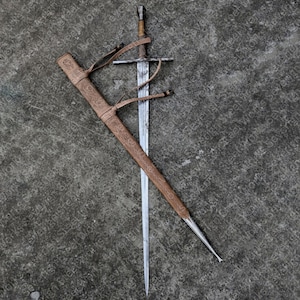 Op de afbeelding: Een middeleeuws zwaard met een zilveren lemmet en een met leer omwikkelde handgreep, naast de bijpassende leren schede. De schede is bruin met reliëfdesigns en leren riemen. Het zwaard is een historisch wapen.