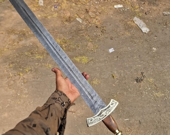 May include: A long, ornate sword with a dark, textured blade. The hilt features a wooden grip, gold accents, and decorative metalwork. The sword is held against a blurred outdoor background.
