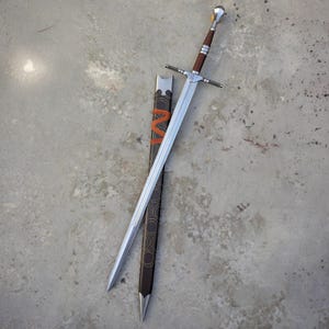 May include: A long steel sword with a double-edged blade, a silver crossguard, and a brown leather-wrapped handle with a silver pommel. Beside it lies a black scabbard featuring a repeating circular pattern and a bright orange cord wrap.