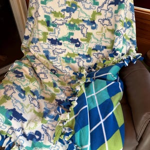 Op de afbeelding: Een omkeerbare fleece deken met een dinosauruspatroon in blauw en groen op een witte achtergrond. De achterkant heeft een blauw, groen en wit ruitpatroon. De randen zijn afgewerkt met geknoopte franjes.