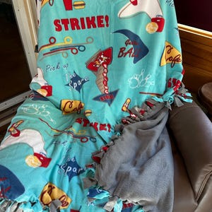 May include: A turquoise fleece blanket with a bowling theme. The blanket features bowling pins, bowling balls, and the word "STRIKE!" in red. The edges are tied with gray, red, and turquoise fabric strips. The blanket is draped over a chair.
