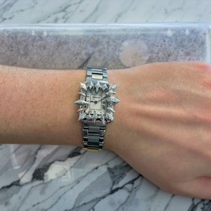 Könnte beinhalten: Eine silberne Uhr mit quadratischem Zifferblatt und silbernem Armband. Die Uhr ist mit kleinen, spitzen silbernen Nieten um das Zifferblatt verziert.