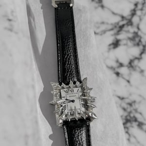 Könnte beinhalten: Eine silberne Uhr mit einem schwarzen Lederarmband. Das Zifferblatt der Uhr hat römische Ziffern und das Wort "SANDOZ" ist darauf gedruckt. Die Uhr ist von einem silbernen Rahmen mit Spikes umgeben.