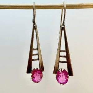 May include: A pair of silver-toned earrings with a geometric design. Each earring features a triangular frame with horizontal bars and a vibrant pink, oval-shaped gemstone at the bottom. The earrings are suspended from a silver hook.